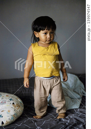 Toddler Standing On Bed, A joyful photo of toddler standing on bed, small child balancing with big smile and outstretched arms, perfect for childhood or family moment documentary. 139974547