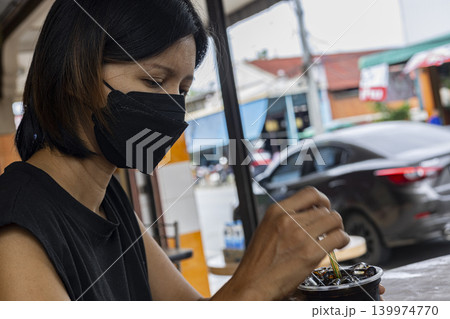 Woman Wearing Face Mask With Coffee. Asian female in black surgical mask holding coffee cup and eating at outdoor cafe, perfect for new normal lifestyle or public health themes. Woman Wearing Face Mask With Coffee. Asian female in black surgical mask holding coffee cup and eating at outdoor cafe, perfect for new normal lifestyle or public health themes. 139974770