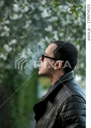 Profile portrait of a young man in a leather jacket smoking outdoors. Cool male character exhaling smoke against a blurred spring blossom background Profile portrait of a young man in a leather jacket smoking outdoors. Cool male character exhaling smoke against a blurred spring blossom background 139974828