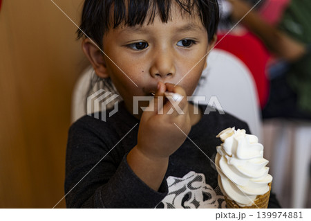 Toddler Boy Sitting And Eating Vanilla Soft Serve Ice Cream. Asian little boy enjoying a sweet dessert cone in a restaurant, perfect for childhood, family lifestyle and summer treat themes. Toddler Boy Sitting And Eating Vanilla Soft Serve Ice Cream. Asian little boy enjoying a sweet dessert cone in a restaurant, perfect for childhood, family lifestyle and summer treat themes. 139974881