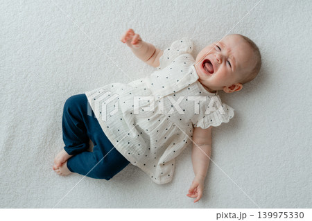 A crying and upset baby girl lies on her back on a soft white blanket. Infant child expressing frustration, discomfort, colic, or hunger during a bright home or studio lifestyle photoshoot A crying and upset baby girl lies on her back on a soft white blanket. Infant child expressing frustration, discomfort, colic, or hunger during a bright home or studio lifestyle photoshoot 139975330