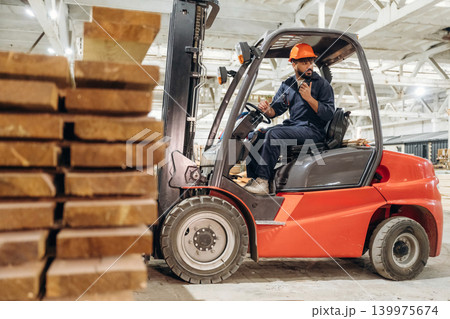 Heavy material, lifting the planks. Warehouse worker operating a forklift while handling wooden materials Heavy material, lifting the planks. Warehouse worker operating a forklift while handling wooden materials 139975674