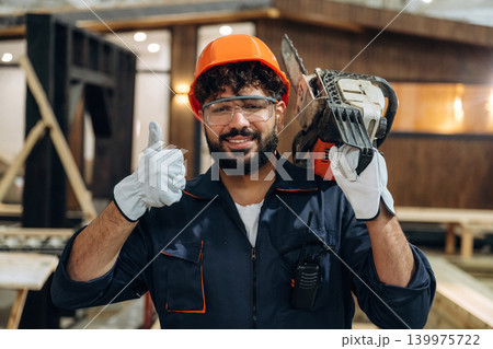 Powerful chainsaw, standing and holding it. Portrait of a male carpenter posing in a woodworking workshop Powerful chainsaw, standing and holding it. Portrait of a male carpenter posing in a woodworking workshop 139975722