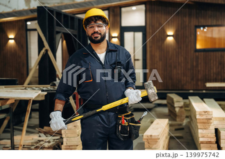 Big hammer is in hands. Portrait of a male carpenter posing in a woodworking workshop Big hammer is in hands. Portrait of a male carpenter posing in a woodworking workshop 139975742