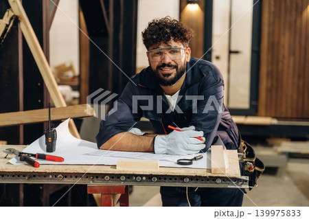 By the table with plan. Portrait of a male carpenter posing in a woodworking workshop By the table with plan. Portrait of a male carpenter posing in a woodworking workshop 139975833