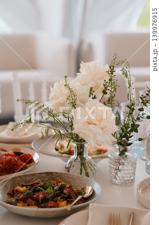 Elegant White Roses in Glass Vase with Gourmet Salad and Appetizers on White Table Elegant White Roses in Glass Vase with Gourmet Salad and Appetizers on White Table 139976915