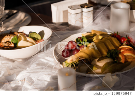 Gourmet Fruit Platter and Cheese Plate on White Linen Tablecloth with Warm Sunlight and Candles 139976947