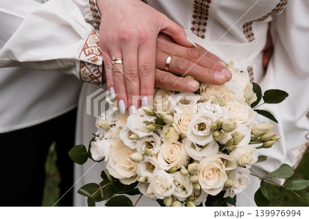 Bride and Groom Hands With Gold Wedding Rings Over White Rose Bouquet in Romantic Wedding Style 139976994
