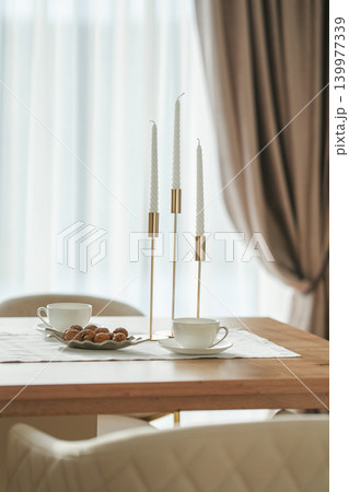 Gold candlesticks with white candles, two elegant white tea and coffee cups and walnuts on a table with a white tablecloth, curtains in the background Gold candlesticks with white candles, two elegant white tea and coffee cups and walnuts on a table with a white tablecloth, curtains in the background 139977339