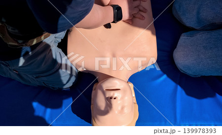 A professional paramedic teacher is teaching a boy first aid and CPR training for kids using a medical mannequin. This outdoor workshop focuses on practicing chest compressions A professional paramedic teacher is teaching a boy first aid and CPR training for kids using a medical mannequin. This outdoor workshop focuses on practicing chest compressions 139978393