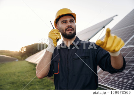 Talking by a walkie talkie. Male solar engineer examining photovoltaic panels at a power station Talking by a walkie talkie. Male solar engineer examining photovoltaic panels at a power station 139978569