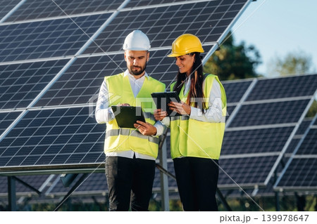 Discussing a plan, showing the area. Two solar engineers standing together at a photovoltaic field, in bright safety vests Discussing a plan, showing the area. Two solar engineers standing together at a photovoltaic field, in bright safety vests 139978647
