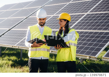 Laptop is in hands if woman. Two solar engineers standing together at a photovoltaic field, in bright safety vests 139978671
