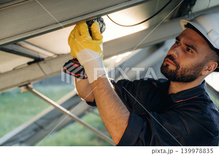 In white hardhat, using drill. Male solar engineer examining photovoltaic panels at a power station In white hardhat, using drill. Male solar engineer examining photovoltaic panels at a power station 139978835