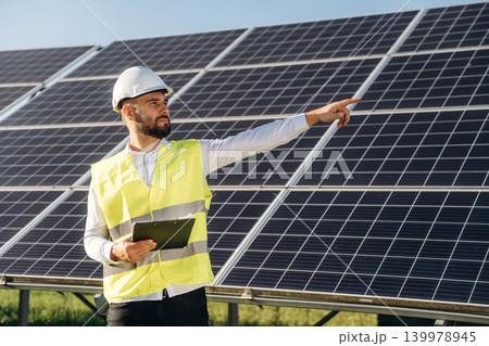 In yellow colored professional uniform. Male solar engineer examining photovoltaic panels at a power station In yellow colored professional uniform. Male solar engineer examining photovoltaic panels at a power station 139978945