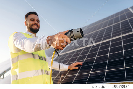 Cable is hand, charging conception. Male solar engineer examining photovoltaic panels at a power station Cable is hand, charging conception. Male solar engineer examining photovoltaic panels at a power station 139978984