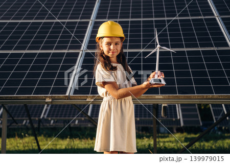 Holding little model of windmill. Happy girl is near renewable energy equipment under bright sunlight 139979015