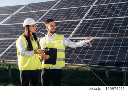 Windmill little model is in hands of woman. Two solar engineers standing together at a photovoltaic field, in bright safety vests Windmill little model is in hands of woman. Two solar engineers standing together at a photovoltaic field, in bright safety vests 139979063