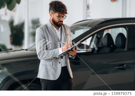 Writing information into notepad. Confident car salesman using digital tablet inside modern showroom 139979375