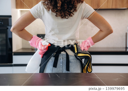 Woman wearing cleaning gloves and apron standing in a modern kitchen, preparing for household chores 139979695