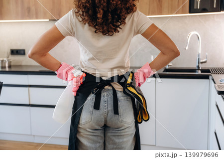 Woman wearing cleaning gloves and apron standing in a modern kitchen, preparing for household chores Woman wearing cleaning gloves and apron standing in a modern kitchen, preparing for household chores 139979696