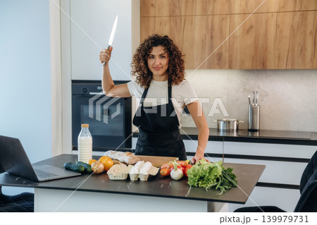Laptop is on the table. Attractive woman preparing fresh vegetables on the kitchen counter during cooking 139979731