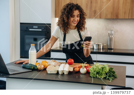 The smartphone is in hands. Attractive woman preparing fresh vegetables on the kitchen counter during cooking The smartphone is in hands. Attractive woman preparing fresh vegetables on the kitchen counter during cooking 139979737