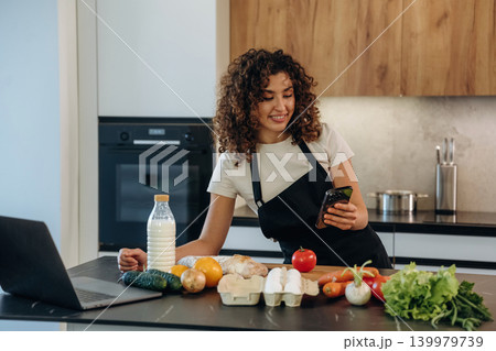 The smartphone is in hands. Attractive woman preparing fresh vegetables on the kitchen counter during cooking The smartphone is in hands. Attractive woman preparing fresh vegetables on the kitchen counter during cooking 139979739