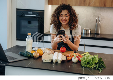 The smartphone is in hands. Attractive woman preparing fresh vegetables on the kitchen counter during cooking The smartphone is in hands. Attractive woman preparing fresh vegetables on the kitchen counter during cooking 139979740