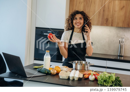 Talking by phone. Attractive woman preparing fresh vegetables on the kitchen counter during cooking Talking by phone. Attractive woman preparing fresh vegetables on the kitchen counter during cooking 139979745