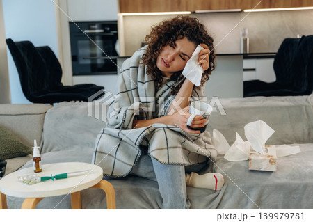 White cup is in hand. Sick woman feeling unwell at home, checking her temperature and using medicine while resting on a sofa White cup is in hand. Sick woman feeling unwell at home, checking her temperature and using medicine while resting on a sofa 139979781