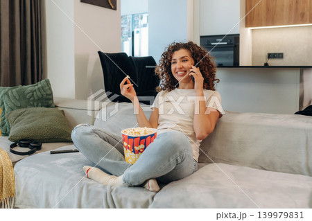Happy woman relaxing on a sofa with popcorn, watching TV and enjoying her leisure time at home 139979831