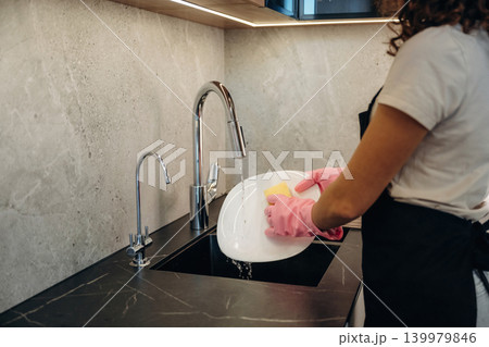 Pink colored gloves. In domestic room. Woman washing dishes with sponge in modern kitchen Pink colored gloves. In domestic room. Woman washing dishes with sponge in modern kitchen 139979846