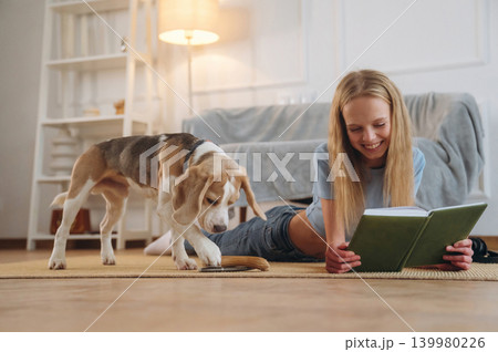 Reading the book in green cover. Happy girl is on the floor at home with dog, domestic room 139980226