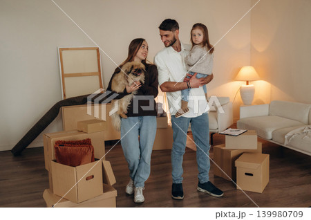 Cute dog is in hands. Happy young family with child moving into new home, standing among cardboard boxes 139980709