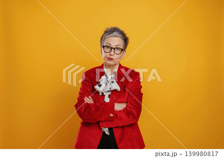 Confident older woman with gray hair and glasses wearing a red blazer, standing with arms crossed against a vibrant yellow background, exuding a strong and assertive presence Confident older woman with gray hair and glasses wearing a red blazer, standing with arms crossed against a vibrant yellow background, exuding a strong and assertive presence 139980817