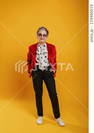 Stylish elderly woman in vibrant red blazer and polka dot blouse poses confidently against a bright yellow background, showcasing modern fashion and self-expression 139980819