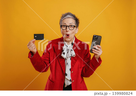 Senior woman in vibrant red blazer excitedly holds smartphone and credit card against bright yellow background, showcasing modern technology and financial empowerment Senior woman in vibrant red blazer excitedly holds smartphone and credit card against bright yellow background, showcasing modern technology and financial empowerment 139980824