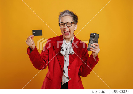 Senior woman in vibrant red blazer joyfully holding smartphone and credit card against bright yellow background, celebrating financial success and modern technology Senior woman in vibrant red blazer joyfully holding smartphone and credit card against bright yellow background, celebrating financial success and modern technology 139980825