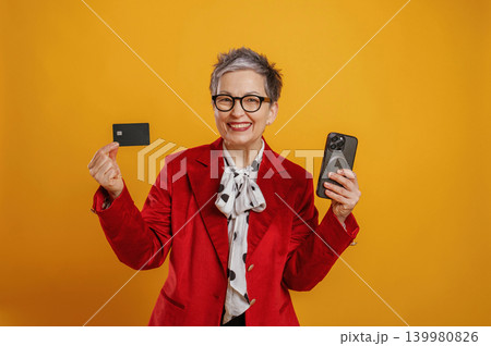 Senior woman in stylish red blazer holding smartphone and credit card, smiling against vibrant yellow background, showcasing modern financial technology and empowerment Senior woman in stylish red blazer holding smartphone and credit card, smiling against vibrant yellow background, showcasing modern financial technology and empowerment 139980826