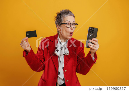 Excited older woman in stylish red blazer holds smartphone and credit card, expressing joy and surprise against vibrant yellow background, showcasing modern technology use Excited older woman in stylish red blazer holds smartphone and credit card, expressing joy and surprise against vibrant yellow background, showcasing modern technology use 139980827