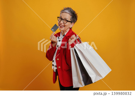 Happy senior woman with stylish glasses holding shopping bags and credit card, celebrating a successful shopping experience against a vibrant yellow background 139980832