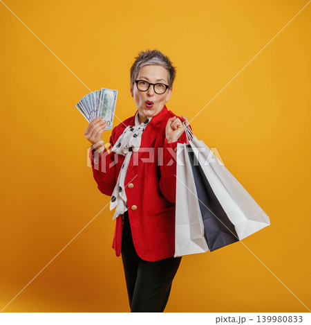 Happy senior woman in stylish outfit holding shopping bags and cash, celebrating a successful shopping spree against a vibrant yellow background Happy senior woman in stylish outfit holding shopping bags and cash, celebrating a successful shopping spree against a vibrant yellow background 139980833