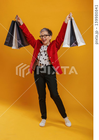 Joyful woman in stylish outfit is celebrating her shopping success while holding colorful bags against a vibrant yellow background, embodying happiness and excitement Joyful woman in stylish outfit is celebrating her shopping success while holding colorful bags against a vibrant yellow background, embodying happiness and excitement 139980841