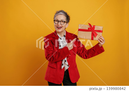 Senior woman in red blazer joyfully holding a gift box with a red ribbon against a vibrant yellow background, celebrating the spirit of giving and joy Senior woman in red blazer joyfully holding a gift box with a red ribbon against a vibrant yellow background, celebrating the spirit of giving and joy 139980842