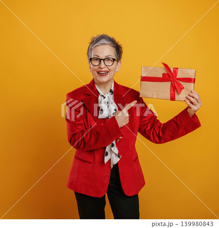 Senior woman in a vibrant red blazer joyfully holding a gift box with a ribbon against a bright yellow background, celebrating the spirit of giving and joy Senior woman in a vibrant red blazer joyfully holding a gift box with a ribbon against a bright yellow background, celebrating the spirit of giving and joy 139980843