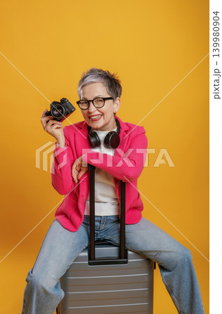 Smiling woman with short gray hair holds camera while sitting on suitcase, wearing pink blazer and headphones, showcasing travel and photography passion Smiling woman with short gray hair holds camera while sitting on suitcase, wearing pink blazer and headphones, showcasing travel and photography passion 139980904