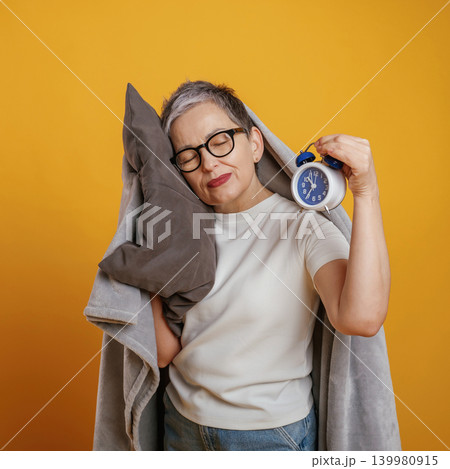 Mature woman with short gray hair embraces a pillow while holding an alarm clock, conveying a sense of relaxation and comfort against a vibrant yellow backdrop Mature woman with short gray hair embraces a pillow while holding an alarm clock, conveying a sense of relaxation and comfort against a vibrant yellow backdrop 139980915