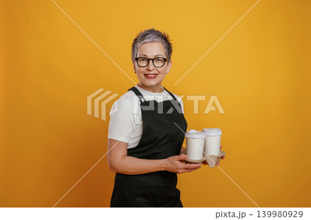 Smiling woman with short gray hair wearing black apron holds coffee cups against vibrant yellow background, showcasing barista skills and customer service Smiling woman with short gray hair wearing black apron holds coffee cups against vibrant yellow background, showcasing barista skills and customer service 139980929