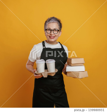 Smiling woman with short gray hair wearing glasses holds a tray with coffee cups and takeout boxes against a vibrant yellow background, showcasing food service and hospitality 139980931
